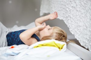 Happy childhood concept. Cute little boy with blonde hair lying on bed in his white cozy bedroom and playing with his hands. Two-year old child wearing pajamas preparing for sleep in nursery
